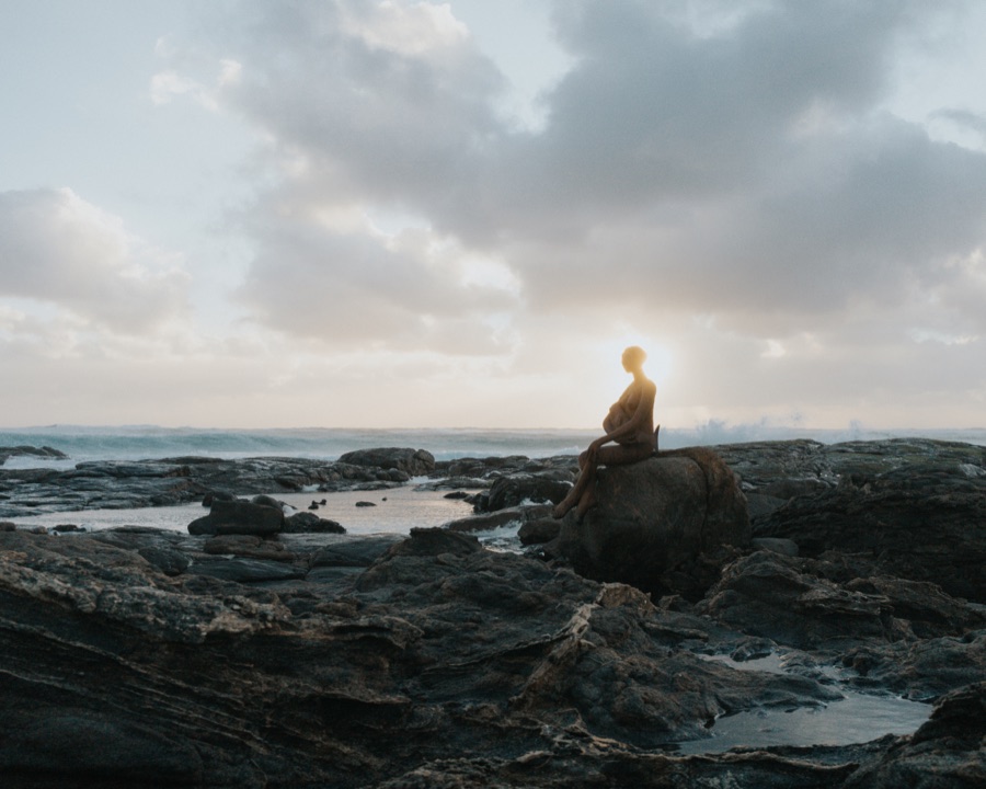 A person seated on a rocky shoreline beneath a wide clouded sky